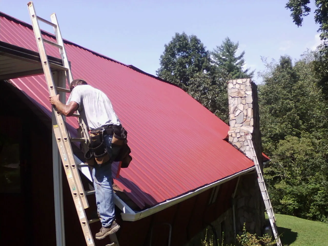 Roofer on a ladder beside a red metal roof and stone chimney — Soffit & Fascia Repair by Roofing Brothers in Lolita